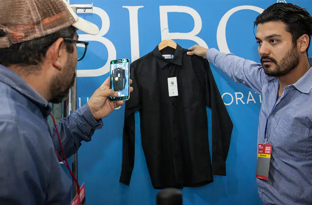 buen-fin-2025-empresas-expositoras Empresario mostrando una prenda a un visitante que la fotografía con su celular, ejemplo de estrategias digitales y exposición de marcas durante el Buen Fin 2025.